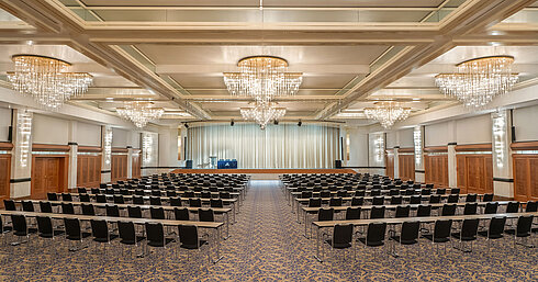 Fürst-von-Metternich-Saal mit großer Bühne, Reihenbestuhlung, eleganten Kronleuchtern und hellem Ambiente.