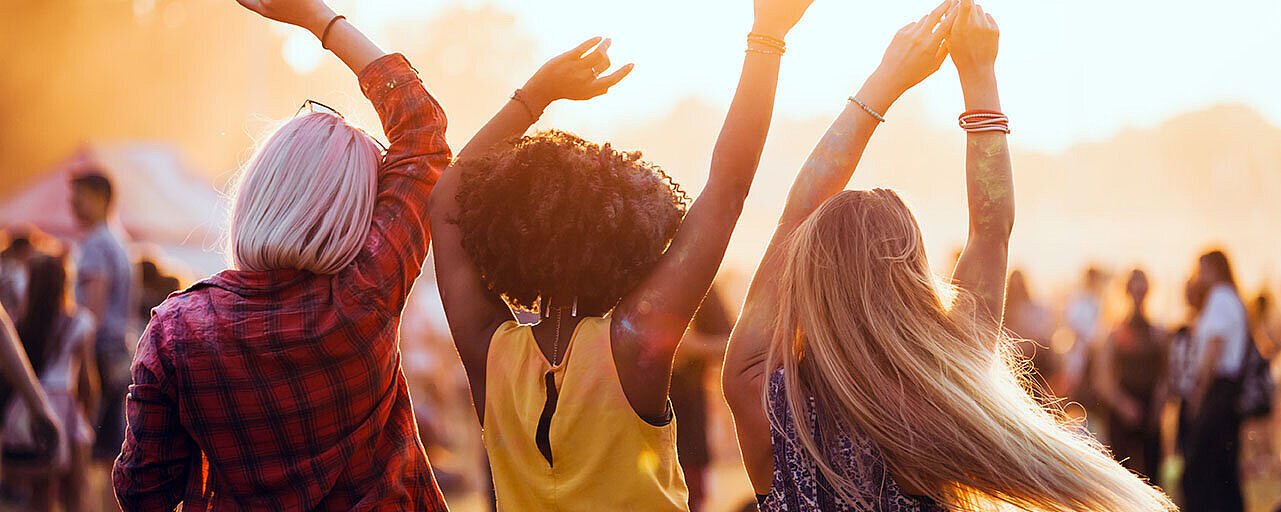 Drei Freunde tanzen mit erhobenen Armen im Sonnenuntergang bei einem Musikfestival im Freien.