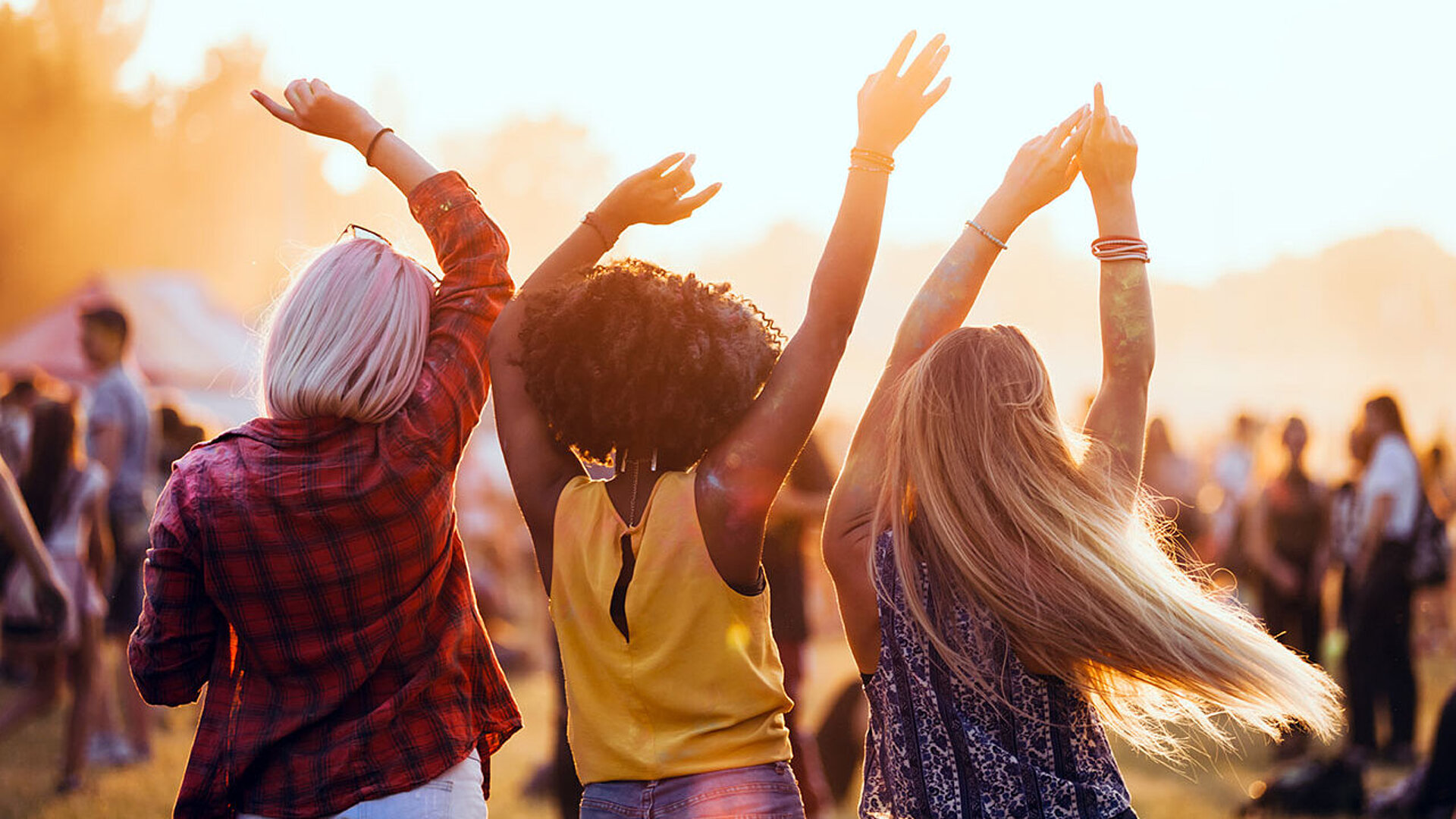 © leszekglasner - AdobeStock.com Drei Freunde tanzen mit erhobenen Armen im Sonnenuntergang bei einem Musikfestival im Freien.