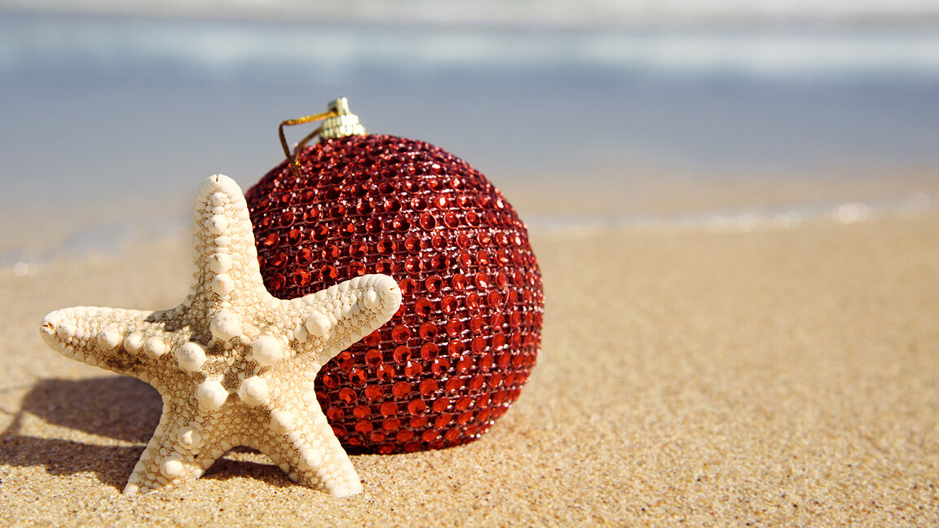 © nito - AdobeStock.com Rote Weihnachtskugel mit Sternmotiv liegt dekorativ im Sand – festliche Stimmung am Strand mit Urlaubsflair
