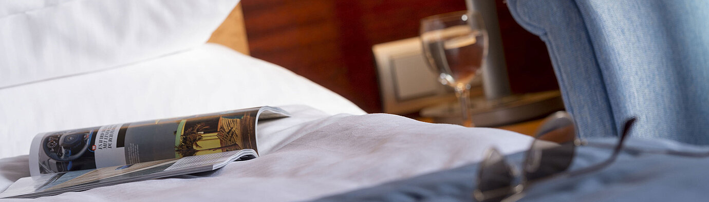 Detailaufnahme eines Hotelzimmers im Maritim Hotel Fulda mit Bett, Zeitschrift, Brille und Glas Wasser auf dem Nachttisch.