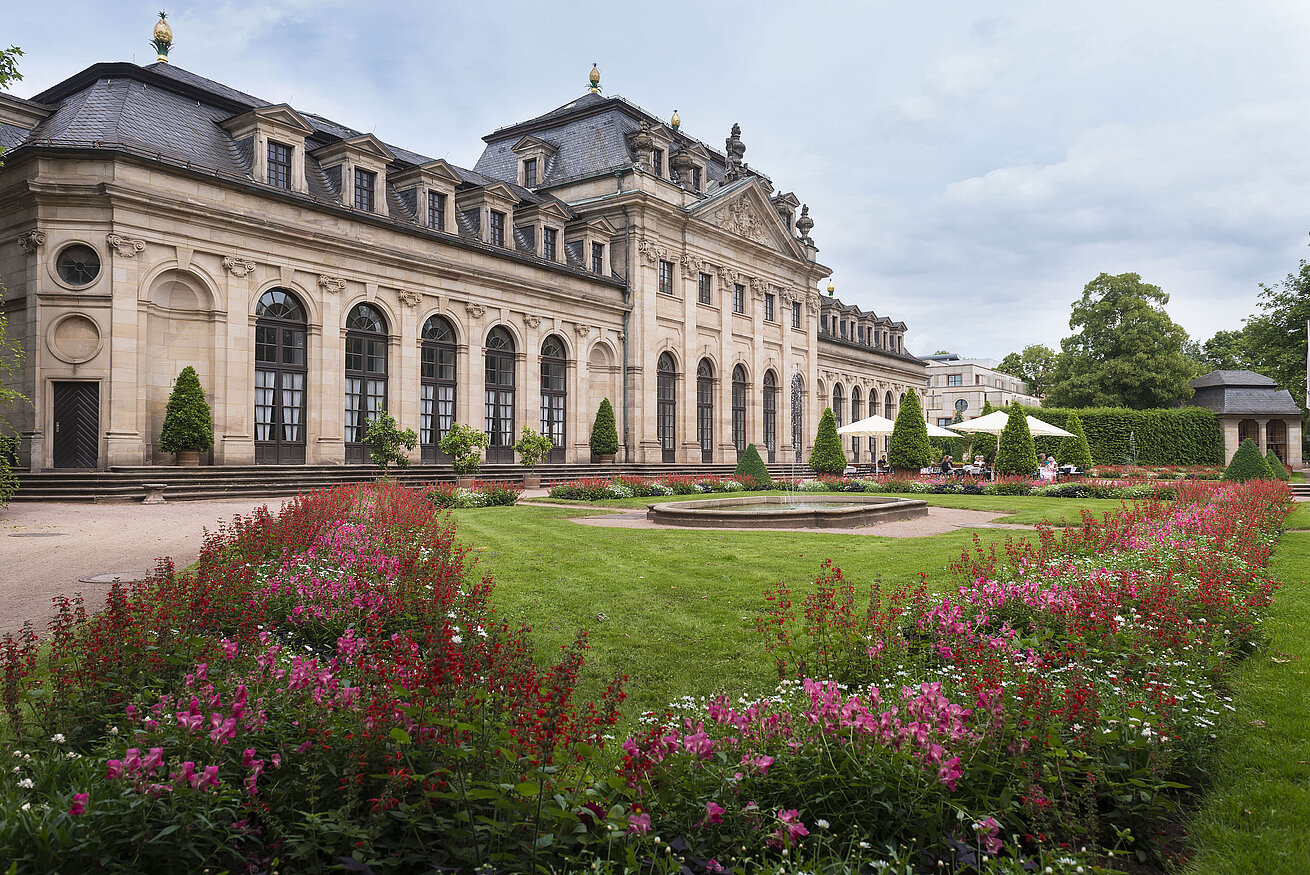 Die historische Orangerie in Fulda mit barocker Fassade, Treppe und blühendem Garten im Vordergrund.