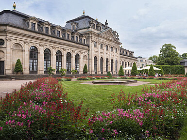 Die historische Orangerie in Fulda mit barocker Fassade, Treppe und blühendem Garten im Vordergrund.