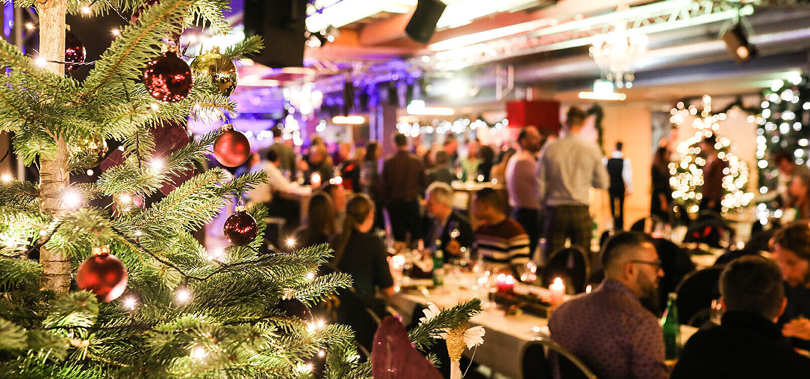 © bilderstöckchen - AdobeStock.com Weihnachtsfeier mit festlich geschmücktem Baum und Gästen im festlichen Saal