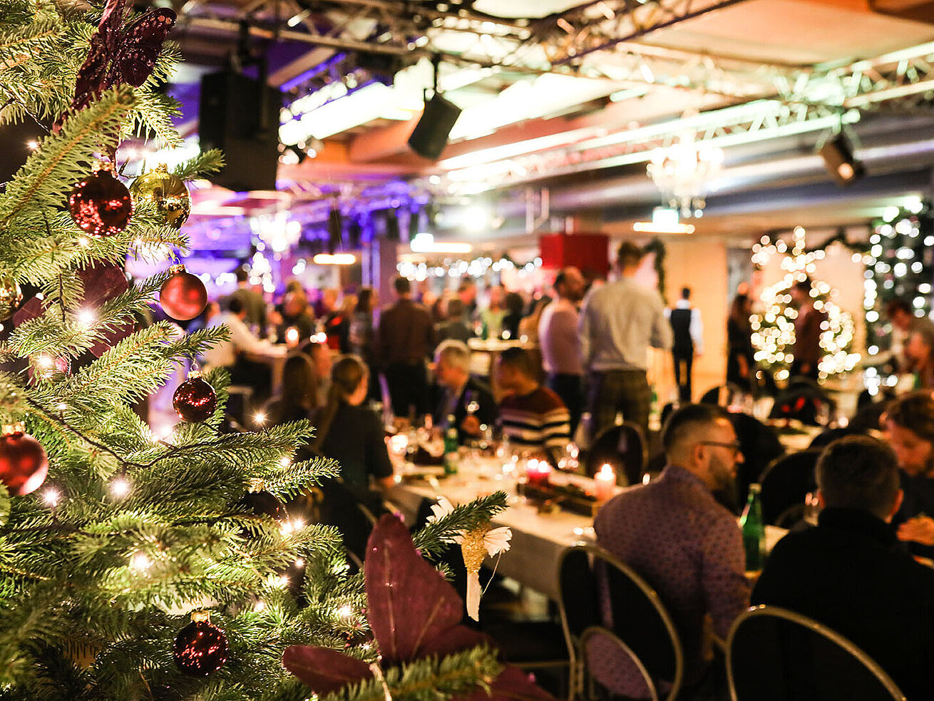 © bilderstöckchen - AdobeStock.com Weihnachtsfeier mit festlich geschmücktem Baum und Gästen im festlichen Saal