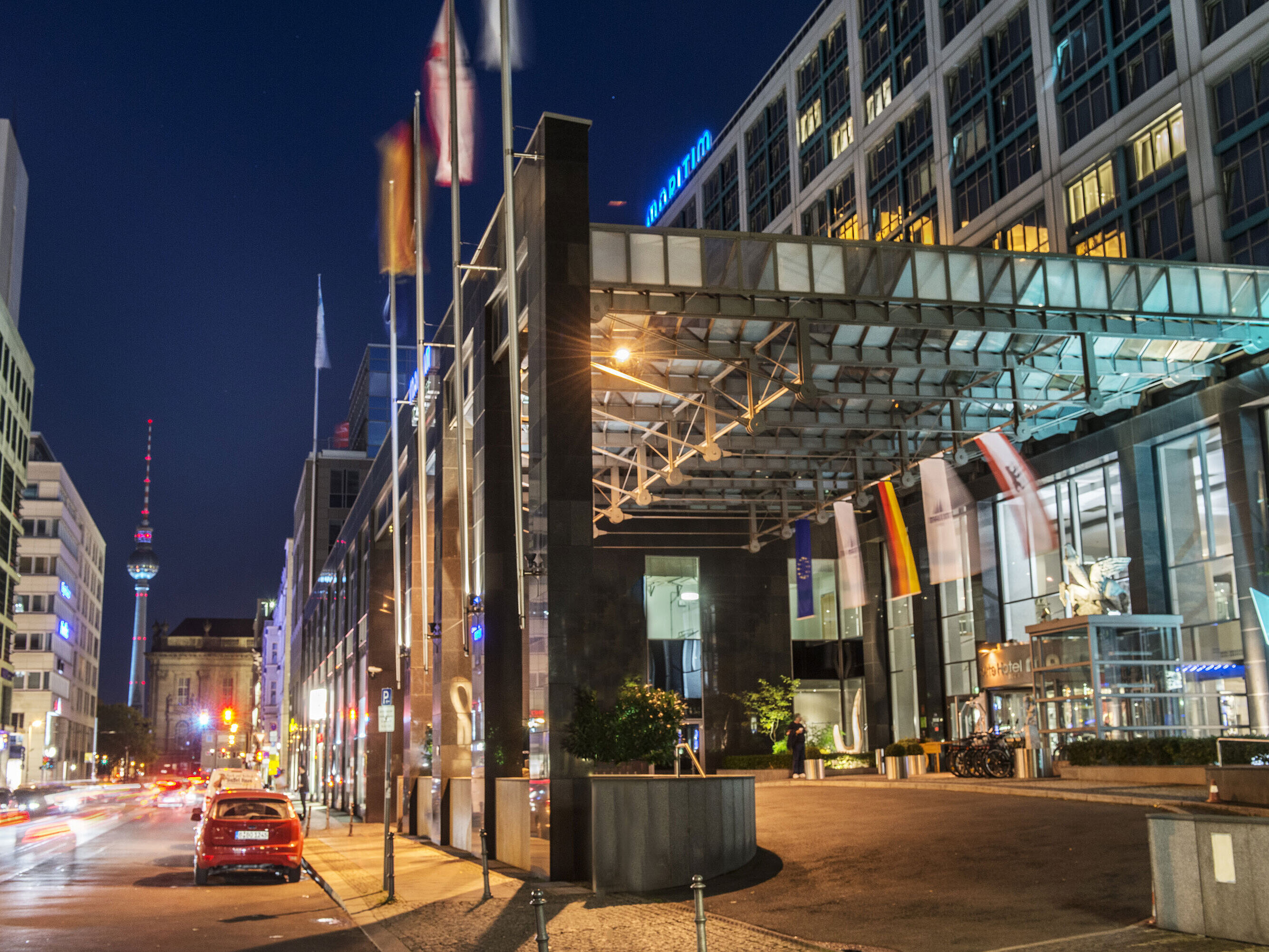 Nachtsicht der beleuchteten Vorfahrt des Maritim proArte Hotels Berlin mit Fernseh-Turm im Hintergrund und Flaggen.