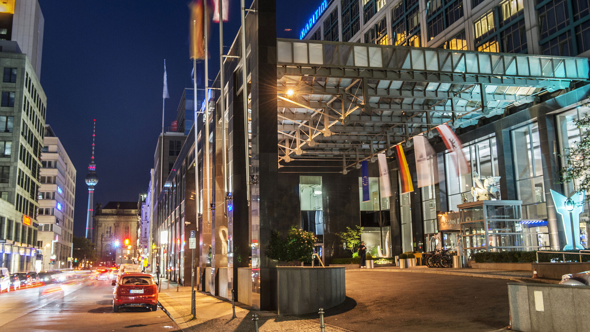 Eingang des Hotels Nachtsicht der beleuchteten Vorfahrt des Maritim proArte Hotels Berlin mit Fernseh-Turm im Hintergrund und Flaggen.