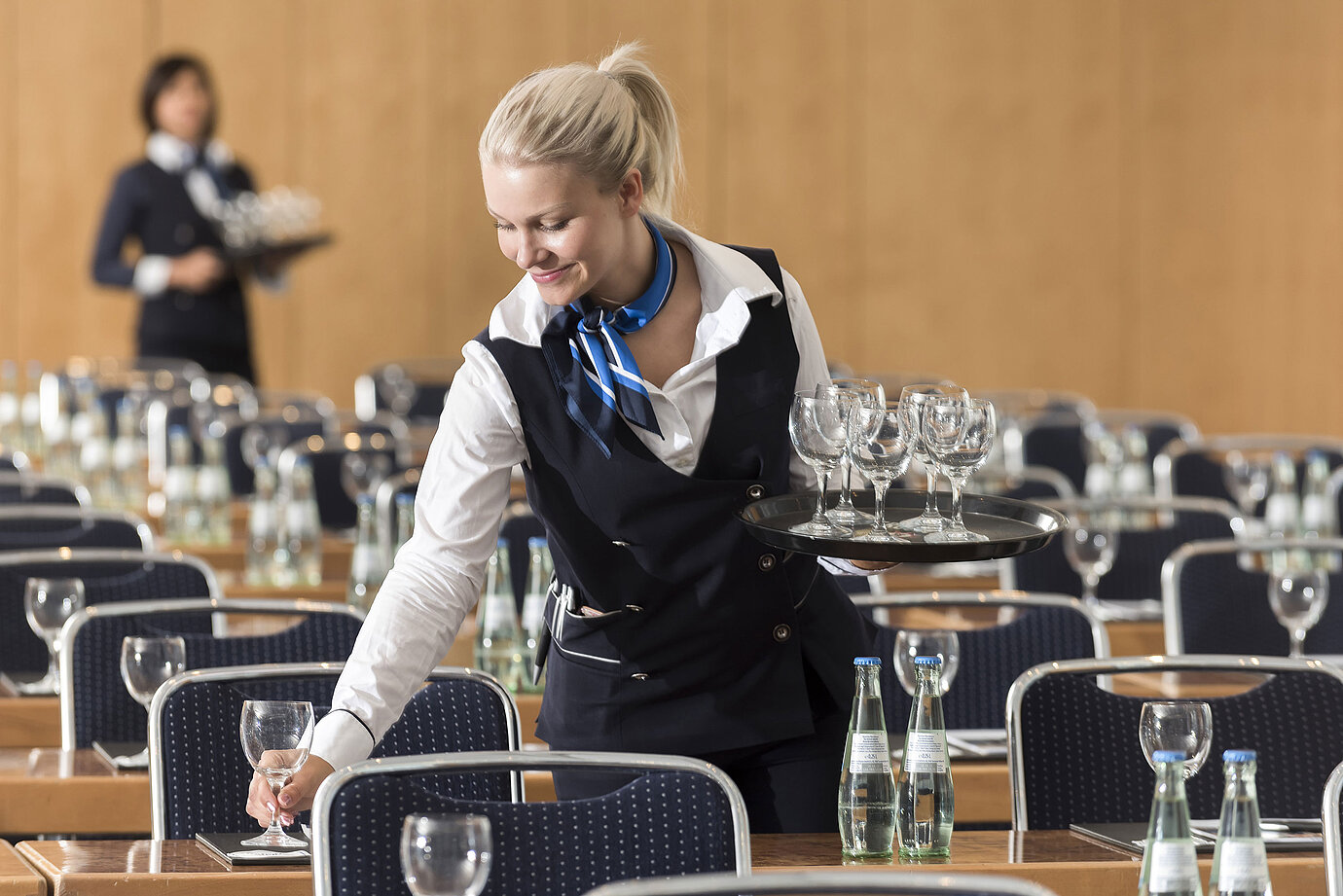 Eine Servicekraft bereitet einen Konferenzraum vor, trägt ein Tablett mit Gläsern und stellt Wasserflaschen bereit.