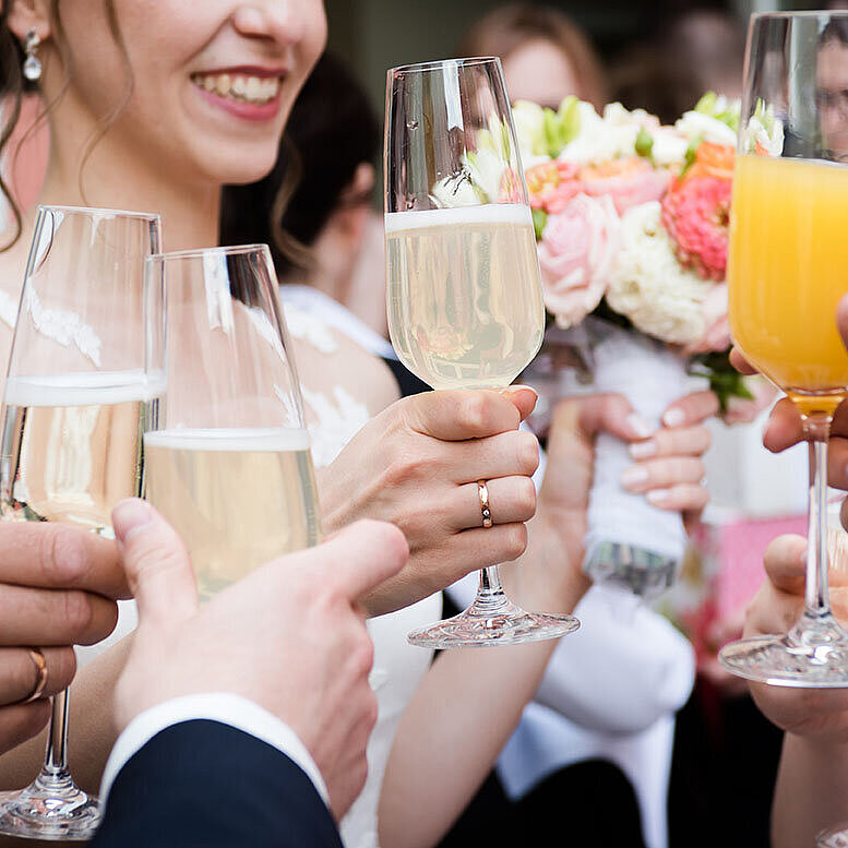 Hochzeitsgäste stoßen mit Sekt und Orangensaft an, während die Braut einen Blumenstrauß hält