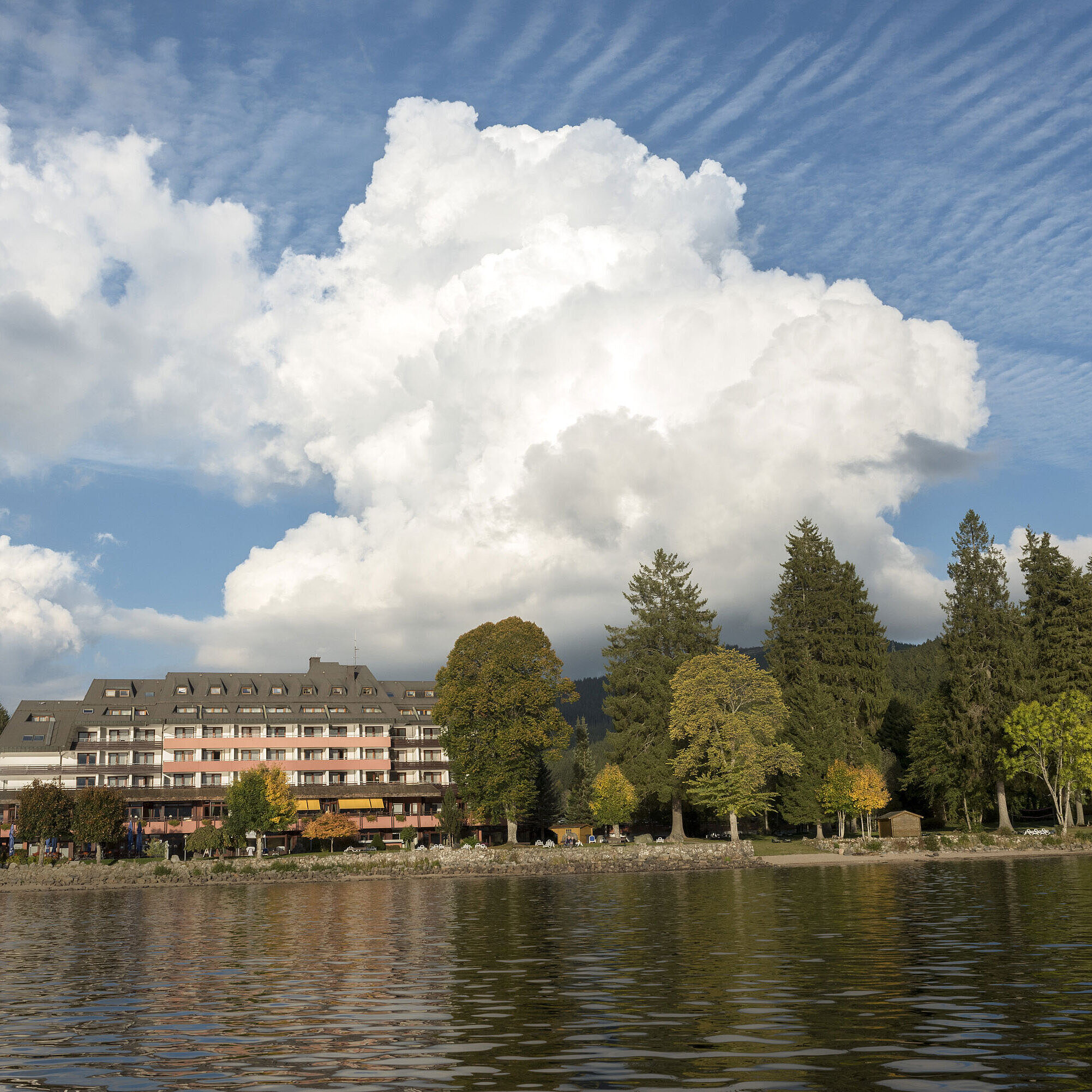 Außenansicht des Maritim Hotel Titisee mit Bäumen und Seeufer bei blauem Himmel und weißen Wolken