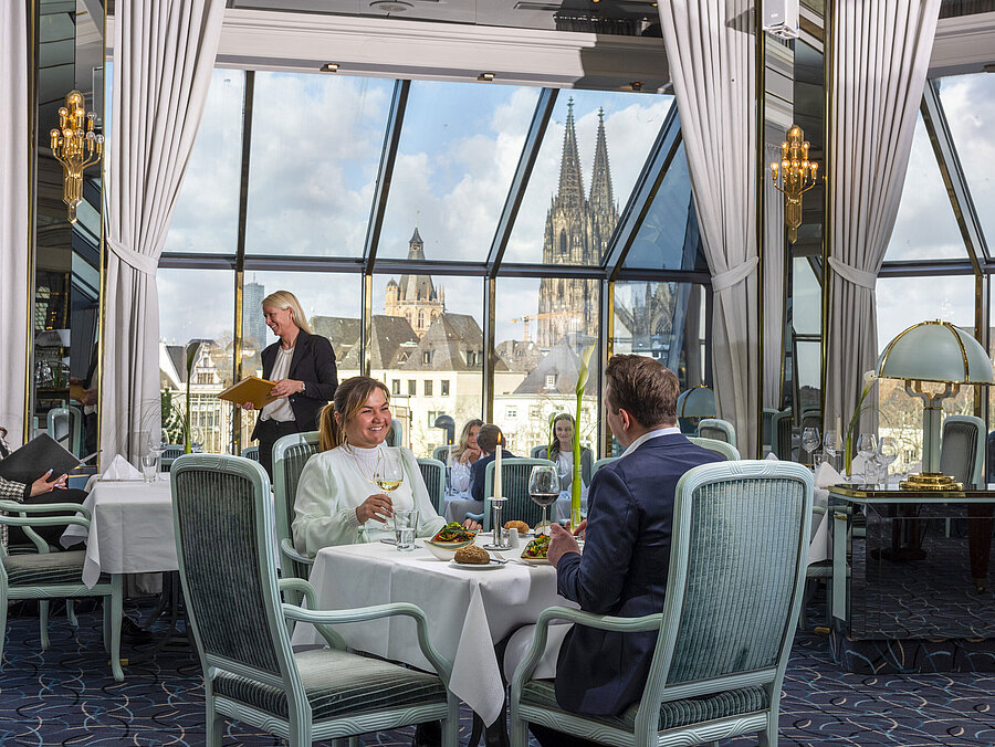 Restaurant "Bellevue" im Maritim Hotel Köln mit Gästen, die speisen, und Blick auf den Kölner Dom durch große Fenster.