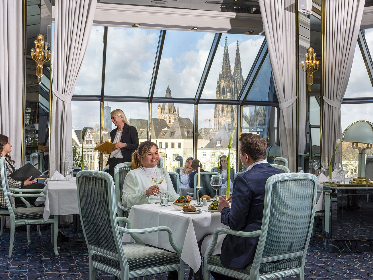 Restaurant "Bellevue" im Maritim Hotel Köln mit Gästen, die speisen, und Blick auf den Kölner Dom durch große Fenster.