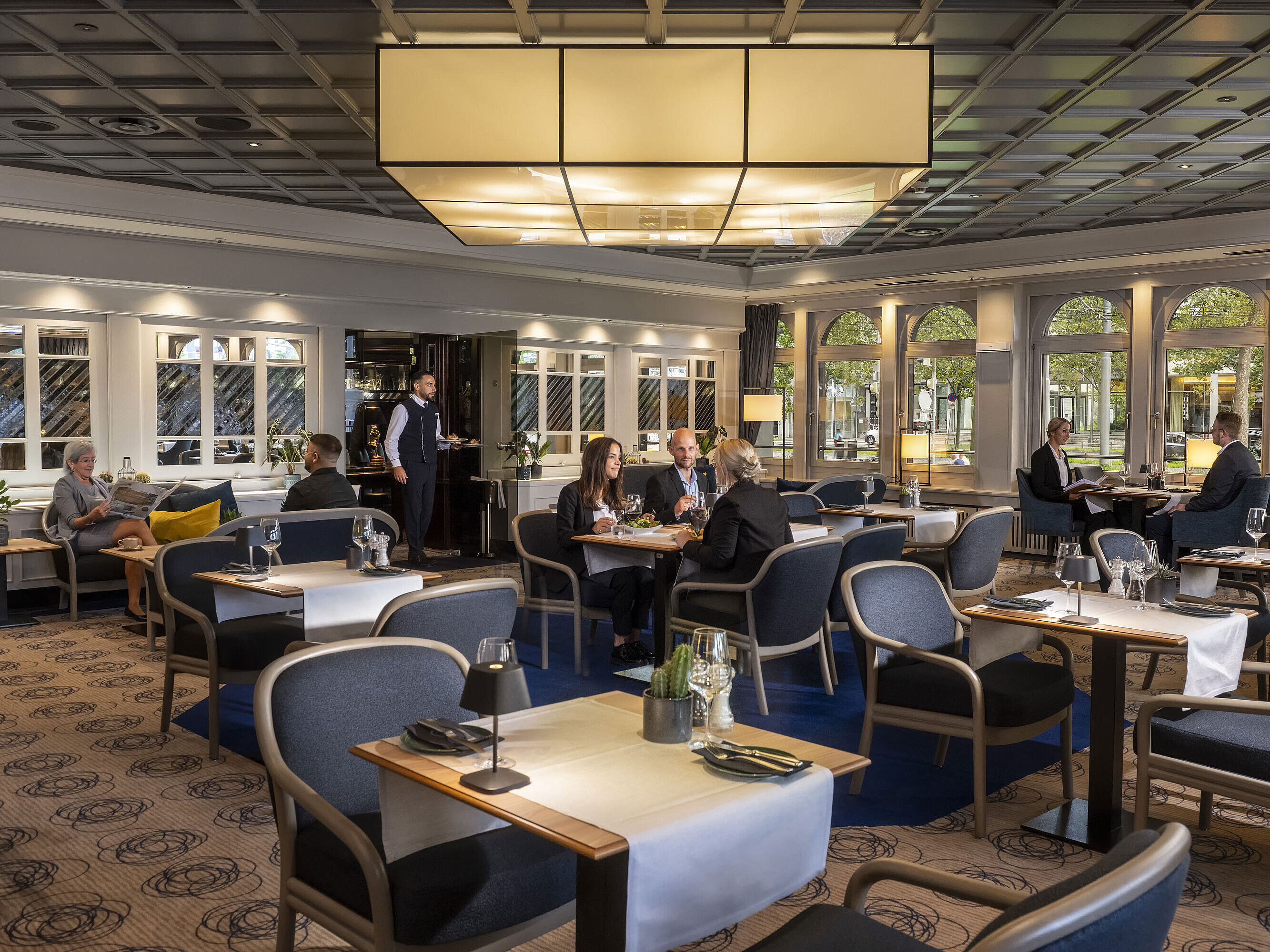 Elegantes Restaurant mit Gästen, die speisen und entspannen. Moderne Einrichtung und große Fenster mit Ausblick.