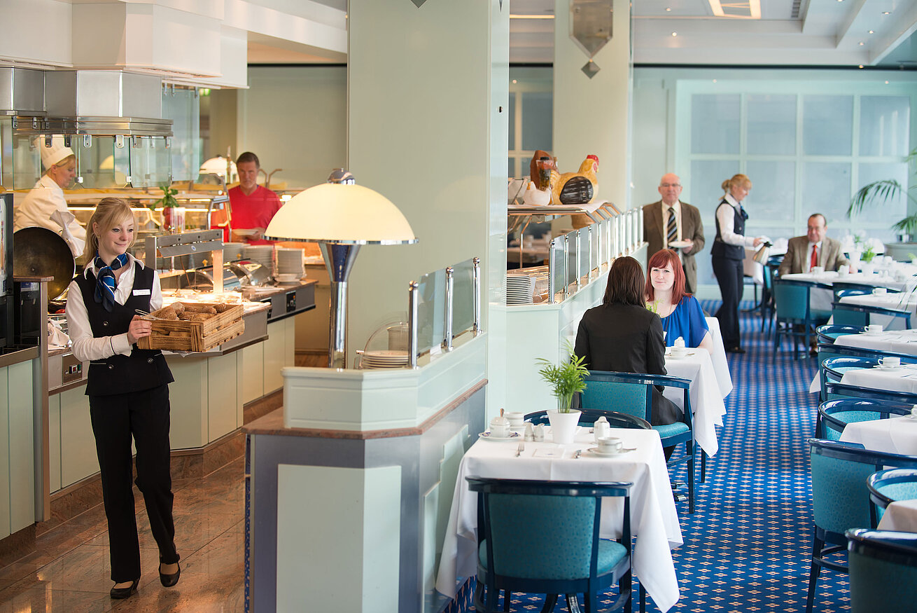 Eine Kellnerin bringt frische Brötchen zum Frühstücksbereich im Maritim Hotel Magdeburg, während Gäste am Tisch sitzen.