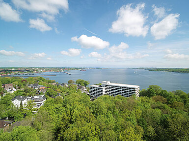 Luftaufnahme des Maritim Hotels Kiel, umgeben von grüner Landschaft und der Kieler Förde, bei strahlend blauem Himmel.