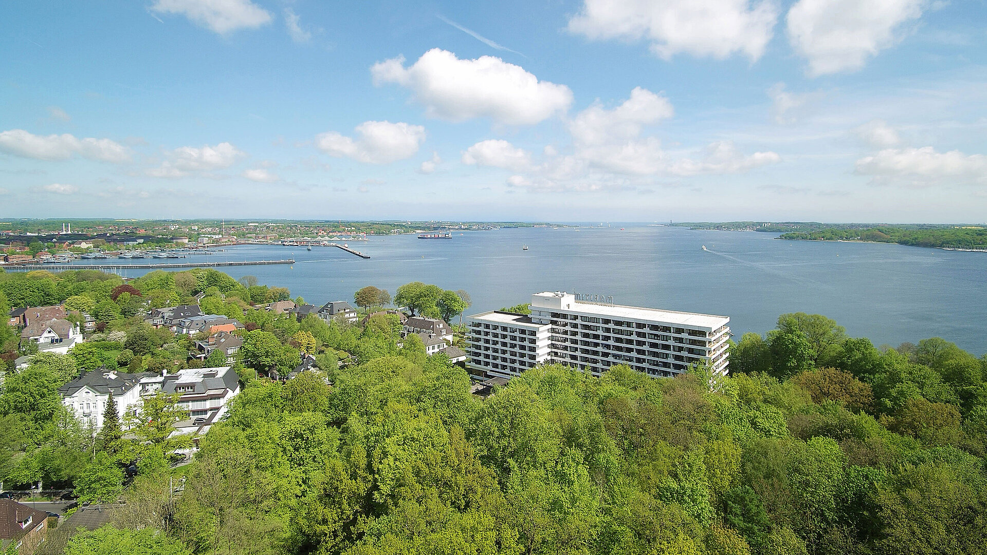Luftaufnahme des Maritim Hotels Kiel, umgeben von grüner Landschaft und der Kieler Förde, bei strahlend blauem Himmel.