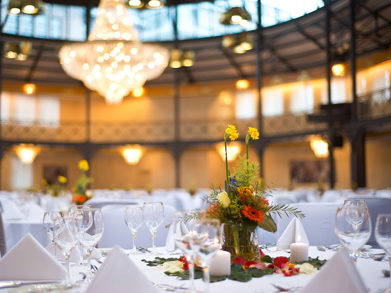 Nahaufnahme einer Tischdekoration mit Blumen und Kerzen, im Hintergrund ein großer Kronleuchter