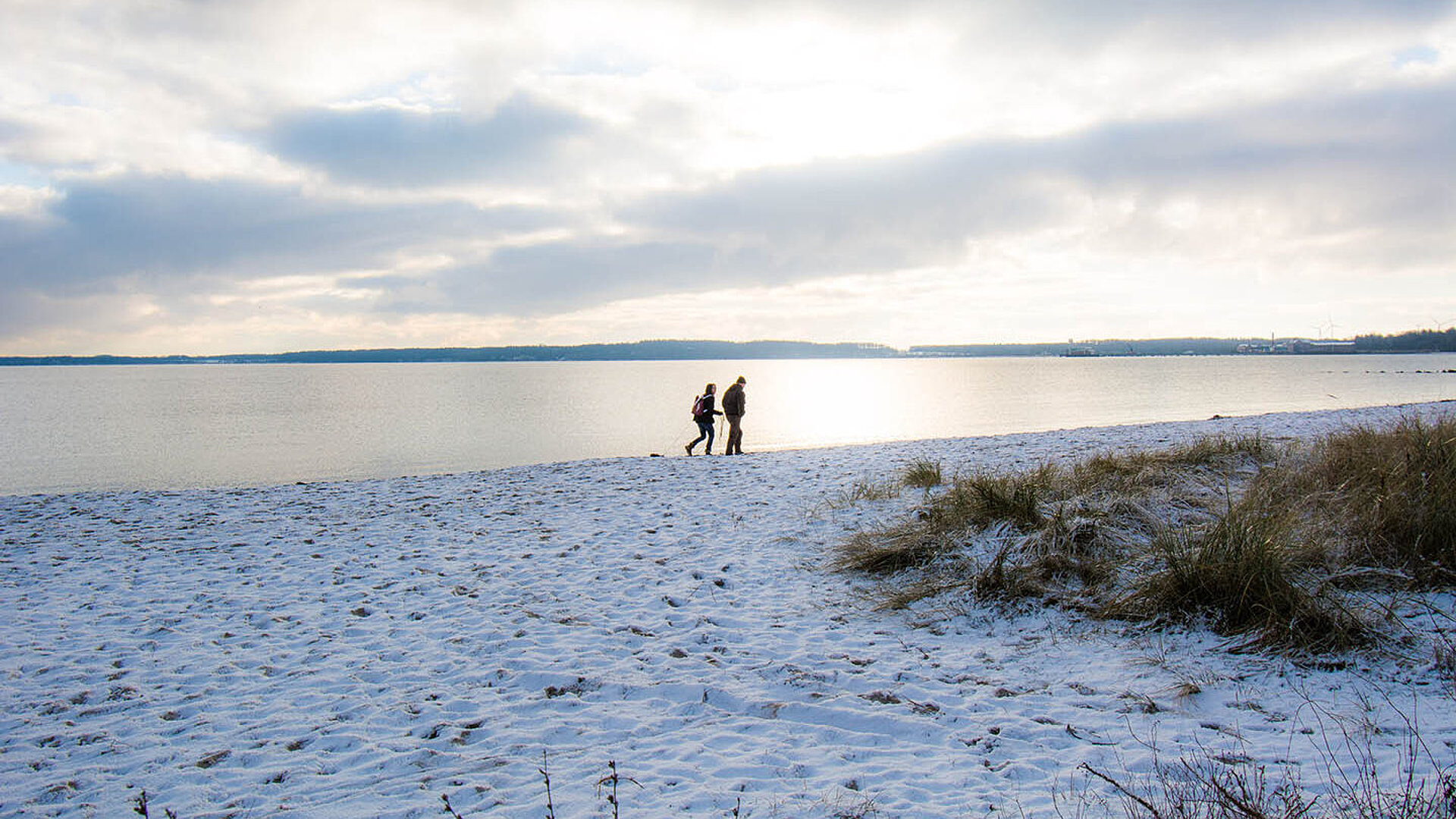 © Stephan - AdobeStock.com Verschneiter Ostseestrand mit zwei Spaziergängern am Wasser unter winterlichem Himmel