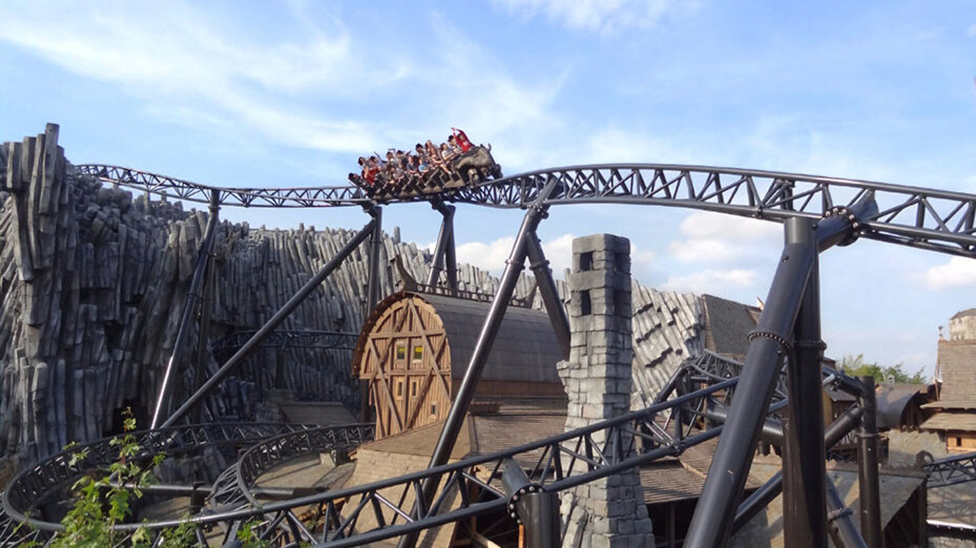 Phantasialand © Danielle - AdobeStock.com Achterbahn Taron in Phantasialand mit Fahrgästen in einer scharfen Kurve zwischen Felsen und thematisierten Gebäuden