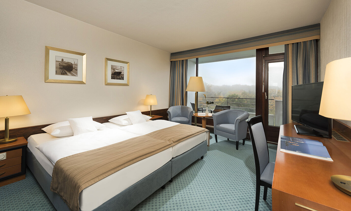 Classic Zimmer im Maritim Hotel Kiel mit großem Doppelbett, Sitzbereich, Schreibtisch und Balkon mit Blick ins Grüne.