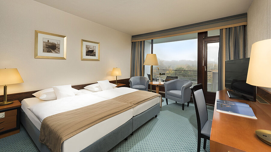 Classic Zimmer im Maritim Hotel Kiel mit großem Doppelbett, Sitzbereich, Schreibtisch und Balkon mit Blick ins Grüne.