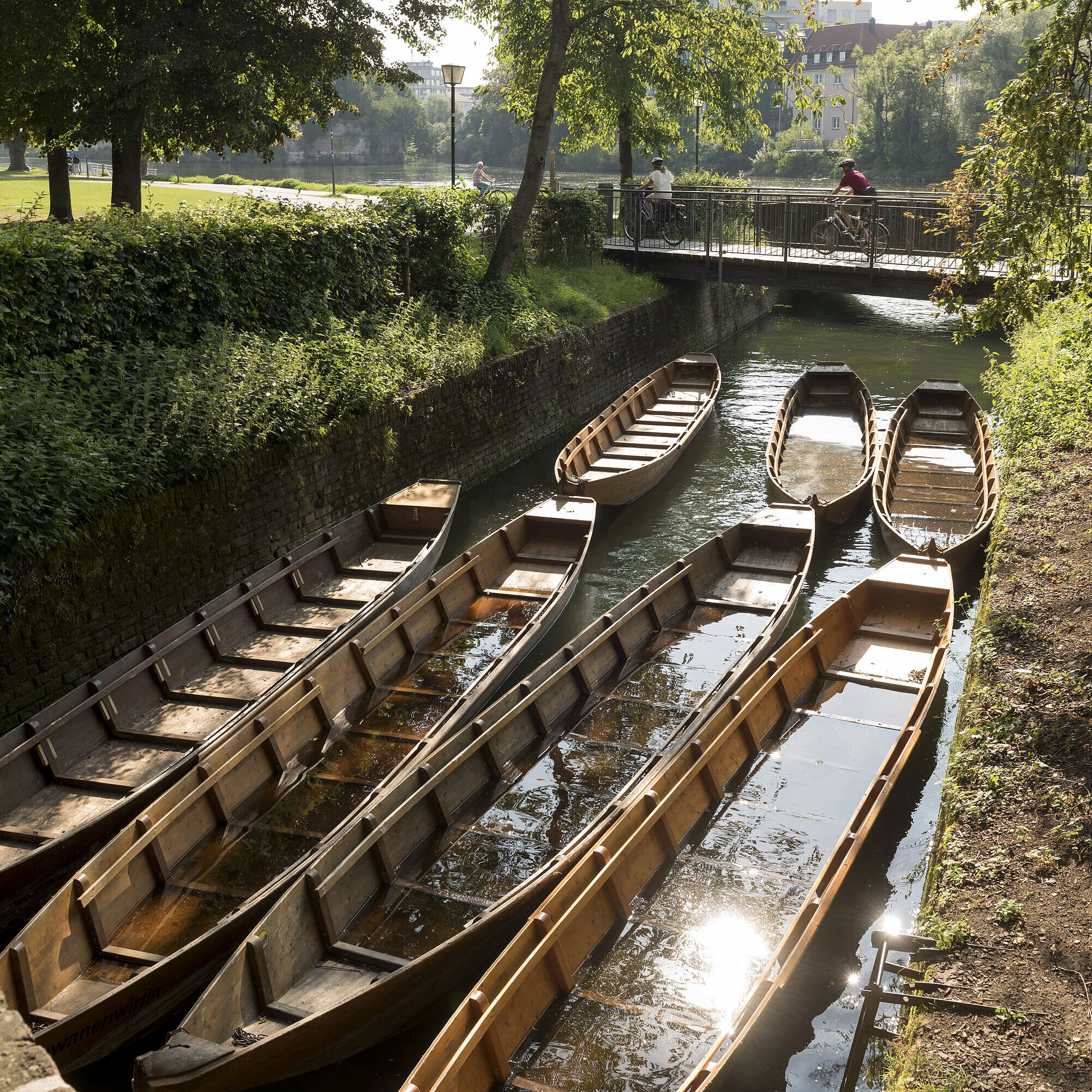 Holzboote in einem Seitenkanal der Donau in Ulm, umgeben von grünen Bäumen.