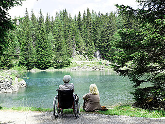 Zwei Personen, eine im Rollstuhl, sitzen am Seeufer und blicken auf den Wald