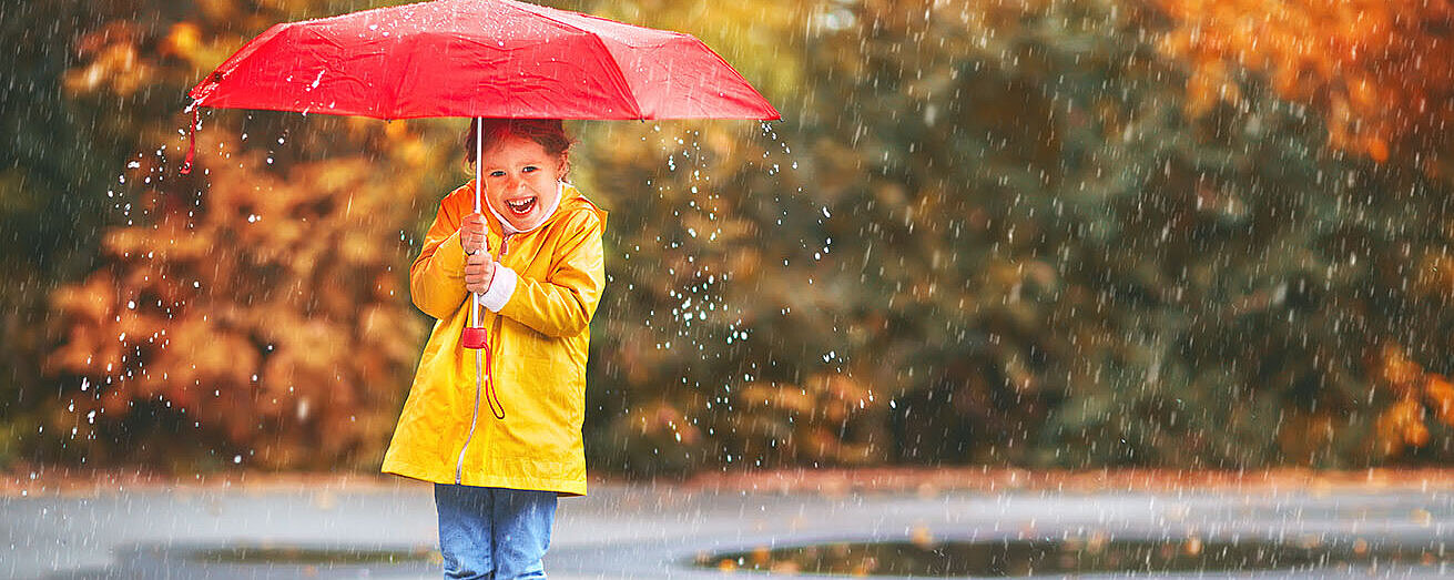 Kind mit gelbem Regenmantel und Stiefeln hält roten Schirm und lacht im herbstlichen Regen.
