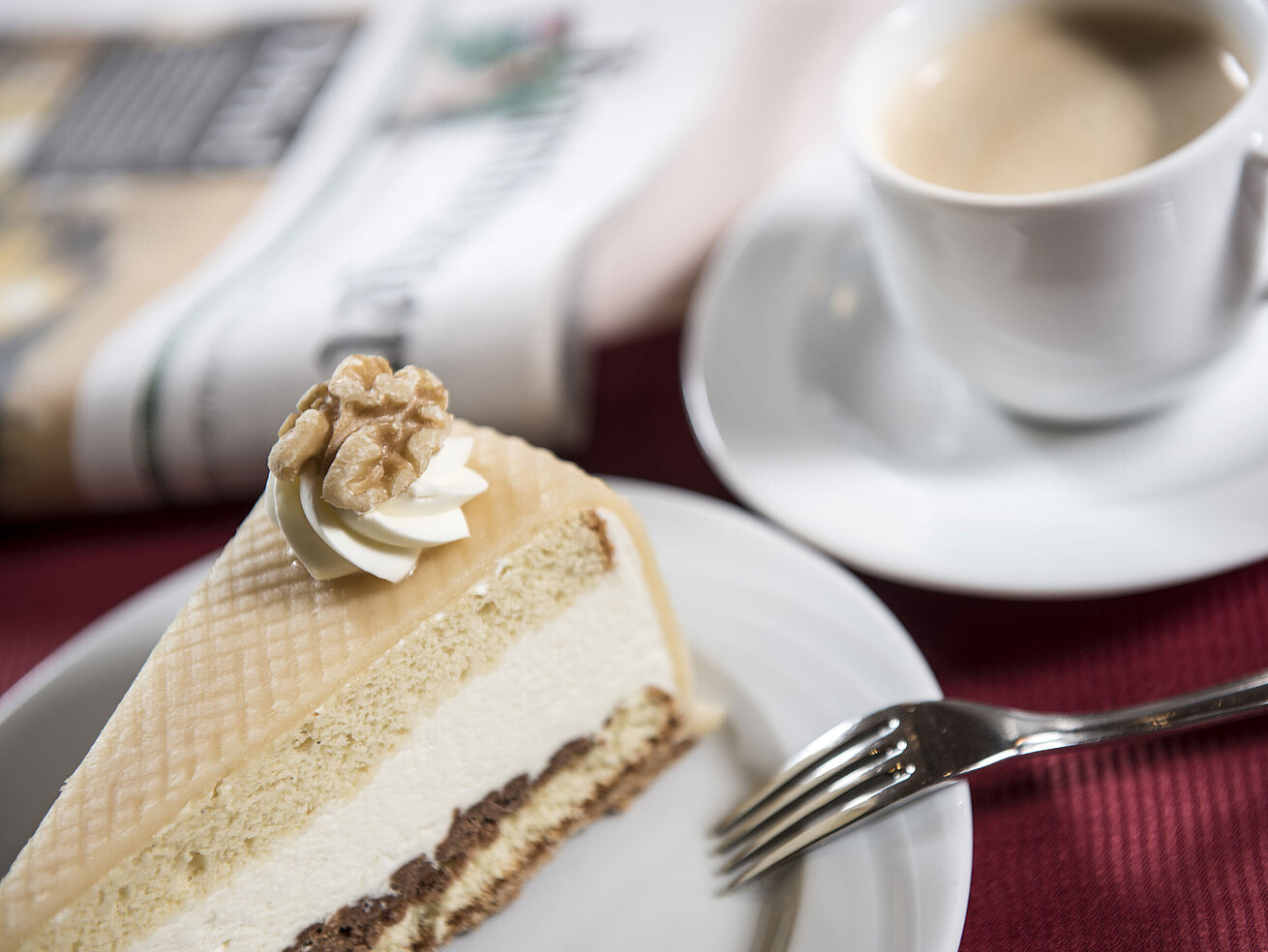 Stück Sahnetorte auf einem Teller mit Gabel, daneben eine Tasse Kaffee und eine Zeitung.