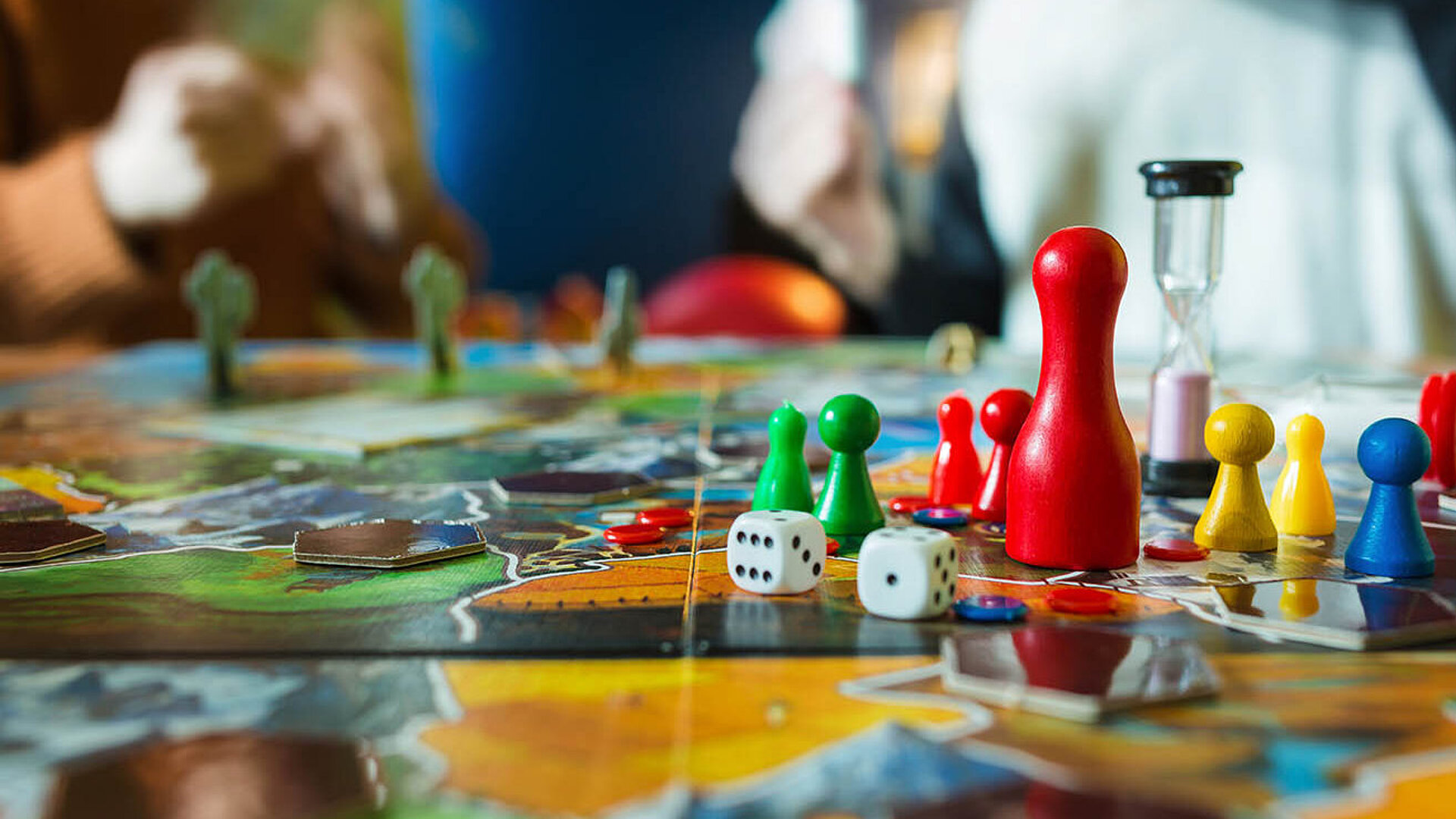 © oleg_ermark-AdobeStock.com.jpg Bunte Spielfiguren, Würfel und Sanduhr auf einem Brettspiel – gemeinsamer Spieleabend mit Familie oder Freunden