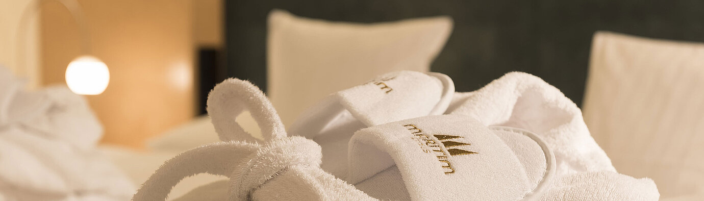 Weißer Bademantel und Hausschuhe mit Maritim-Logo liegen bereit auf dem Bett für entspannte Wellnessmomente im Hotelzimmer
