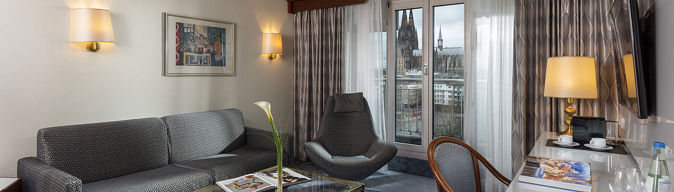 Junior Suite im Maritim Hotel Köln mit Blick auf den Kölner Dom, stilvollem Sofa, Sessel und Schreibtisch mit Lesematerial.