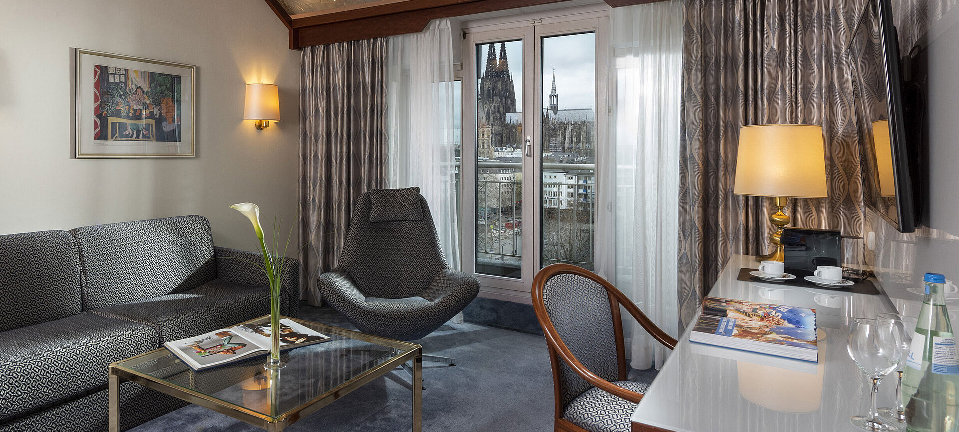 Junior Suite im Maritim Hotel Köln mit Blick auf den Kölner Dom, stilvollem Sofa, Sessel und Schreibtisch mit Lesematerial.