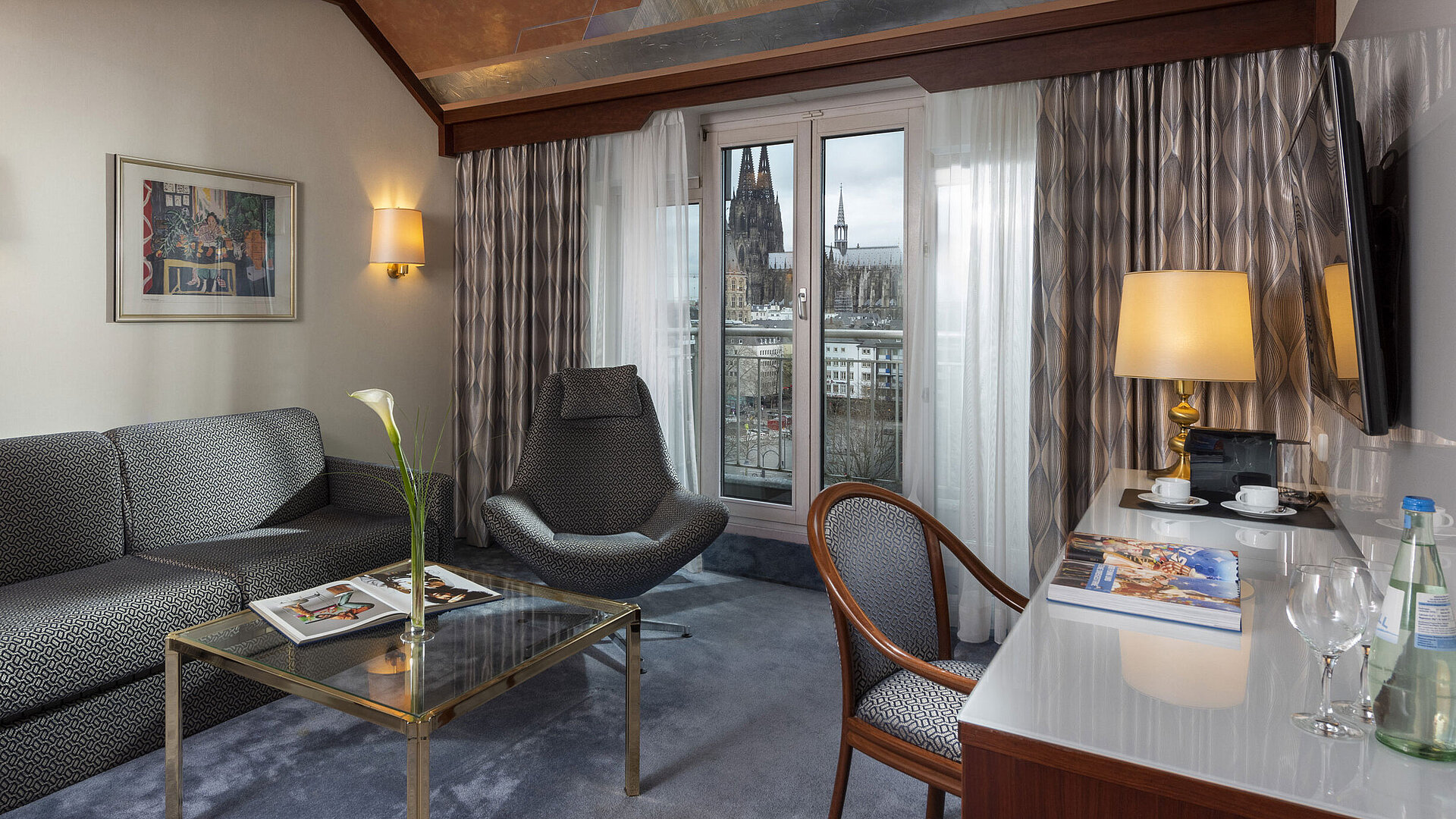 Junior Suite im Maritim Hotel Köln mit Blick auf den Kölner Dom, stilvollem Sofa, Sessel und Schreibtisch mit Lesematerial.