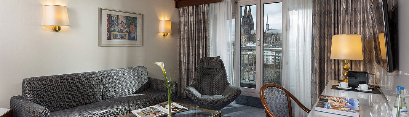 Junior Suite Junior Suite im Maritim Hotel Köln mit Blick auf den Kölner Dom, stilvollem Sofa, Sessel und Schreibtisch mit Lesematerial.