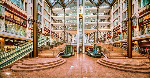 Die geräumige Lobby des Maritim Hotels Köln mit Treppen, Rolltreppen, Glasdach und Geschäften auf mehreren Ebenen.