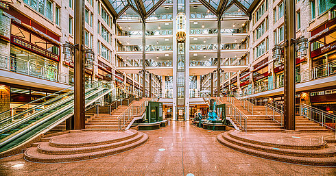 Die geräumige Lobby des Maritim Hotels Köln mit Treppen, Rolltreppen, Glasdach und Geschäften auf mehreren Ebenen.