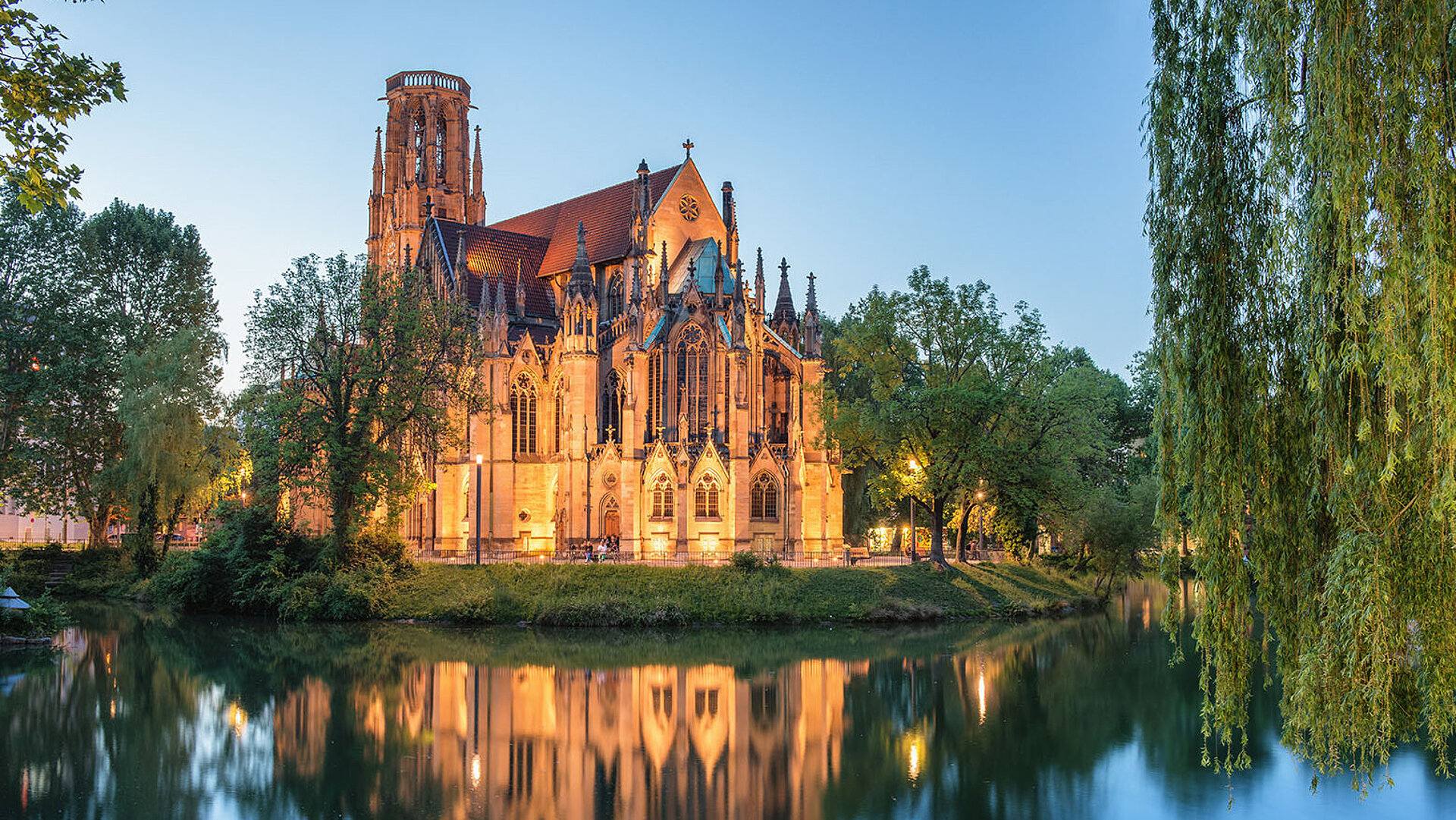 Johanneskirche am Feuersee in Stuttgart mit beleuchteter neugotischer Fassade und Spiegelung im Wasser