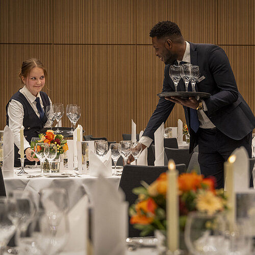Servicepersonal deckt Tische im Saal Barbarossa des Maritim Hotel Würzburg mit Blumenschmuck und Gläsern ein.