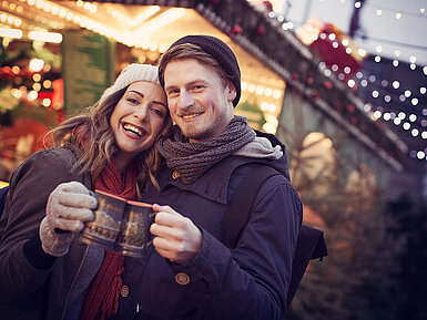 © hailey copter - AdobeStock.com Paar stößt mit Glühwein auf einem festlich beleuchteten Weihnachtsmarkt an