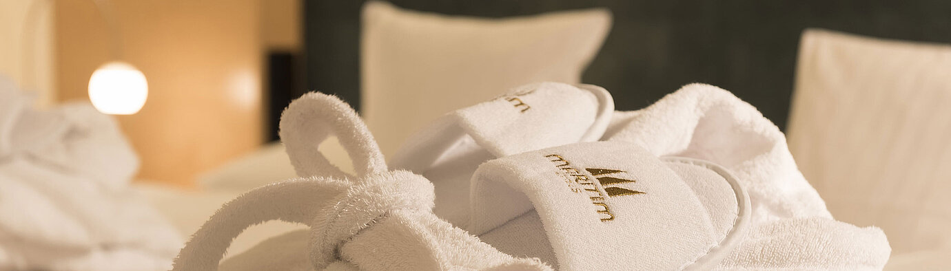 Bademantel  Ein flauschiger Bademantel und Hausschuhe mit Maritim-Logo auf einem Bett im Maritim proArte Hotel Berlin.