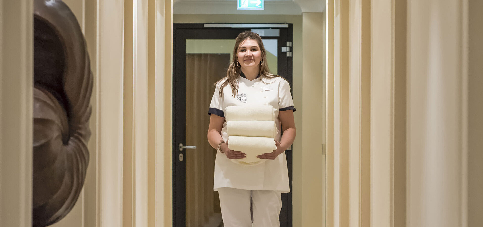 Wellness-Mitarbeiterin im Spa-Bereich des Maritim Hotels, die Handtücher in einem stilvollen Korridor trägt.