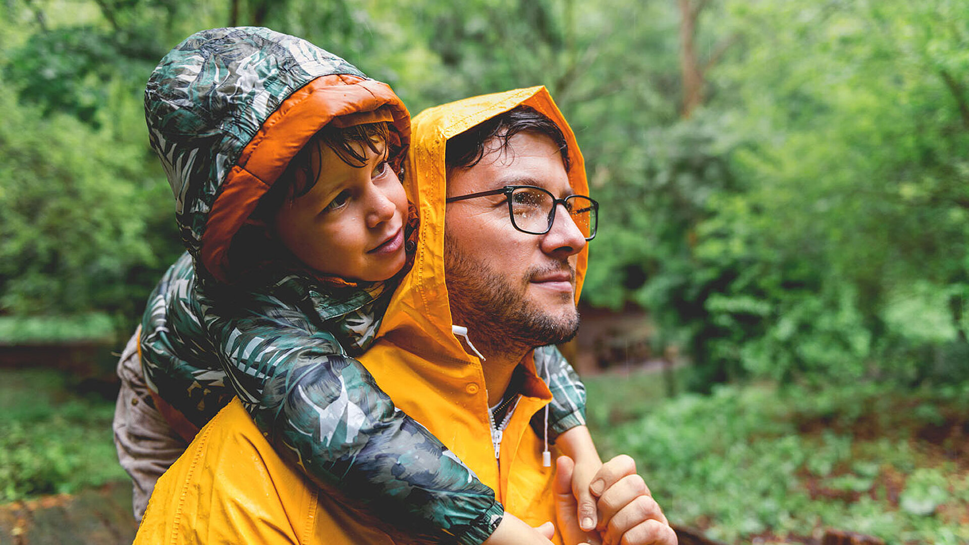Erwachsene Person trägt ein Kind im Regen durch einen grünen Wald, beide mit Regenjacken.