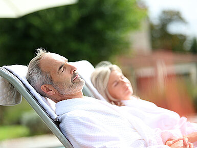 © goodluz - AdobeStock.com Mann und Frau im Bademantel entspannen auf Liegen im Freien und genießen eine erholsame Auszeit.